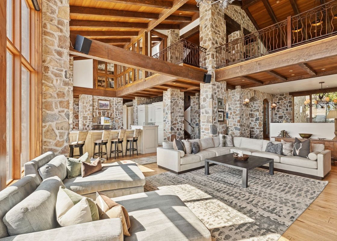 The great room of Snow Pine Lodge at Deer Valley, featuring two large sectional sofas, a dark wood coffee table, a bar area, and rustic stone and wood beam architecture.