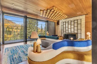 The living area of Monitors Rest features a vibrant, multi-layered sofa, a distinctive gold-toned chandelier, a modern fireplace, and expansive windows with breathtaking views of an autumnal forest.