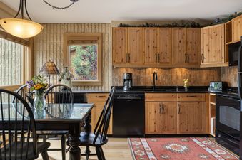 A cozy kitchen and dining area with natural wood cabinets, black appliances, a dark wood dining table with a glass top, and large windows offering views of lush greenery.