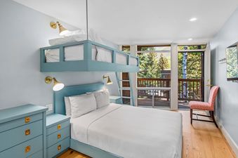 A bedroom with a bunk bed, a large window with a view of the trees, and a bedside table with a lamp.