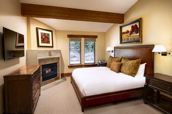The primary bedroom features a large, comfortable bed with an elegant upholstered headboard, a fireplace, a TV, and wooden furnishings.