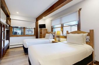 A cozy bedroom at Slope 101 features two twin beds with white linens, a wooden dresser with a TV, and a large window with blinds, all framed by rustic wooden beams.