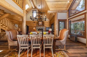 A spacious dining area in Pine Ridge Cabin, featuring a large wooden table set for a meal, surrounded by upholstered chairs, with a cozy fireplace visible in the background and large windows offering a view of the outdoors.