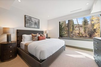 A luxurious bedroom at Riverfront Residences 02 features a queen-sized bed, elegant nightstand, and a large window overlooking a scenic forest.