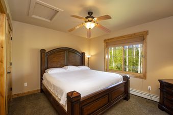 A cozy bedroom with a large wooden bed, a ceiling fan, and a window with a view of lush green trees.