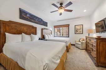 A bedroom at The Charter D3205 features two queen beds with wooden headboards, white bedding, a large mountain landscape painting, and a window with wooden blinds.
