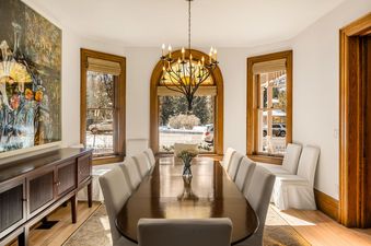 An elegant dining room with a large wooden oval table surrounded by white chairs, illuminated by an ornate chandelier and natural light from several windows showcasing a snowy outdoor view.