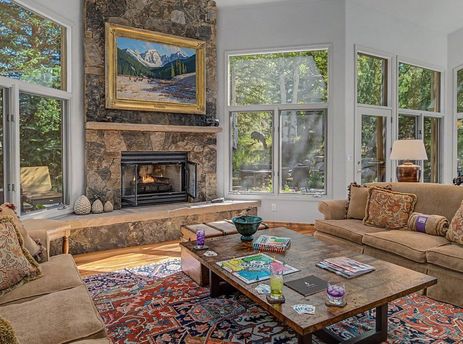 Inviting living room at the Wild Iris Lodge with a stone fireplace, large windows overlooking green woods, and cozy seating around a coffee table.