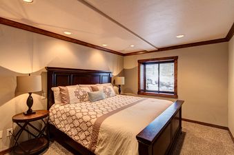 A bedroom with a large bed, a window with blinds, and a side table with a lamp