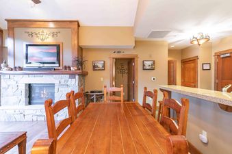 A wooden dining table with chairs sits in the middle of a room with a fireplace, TV, and a door.