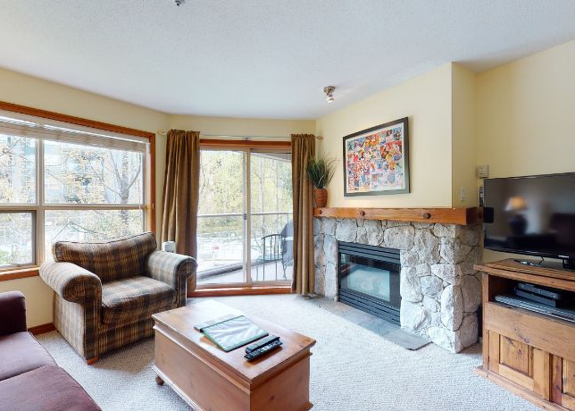 The inviting living area of Aspens on Blackcomb Unit 206 features a fireplace, comfortable seating, and a large television, with a view of the outdoors through the windows.