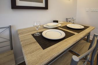 A cozy dining area at Fraser Crossing Studio 3518 with a light wood-toned table set for two, featuring plates, bowls, and wine glasses, accompanied by metal-framed chairs.