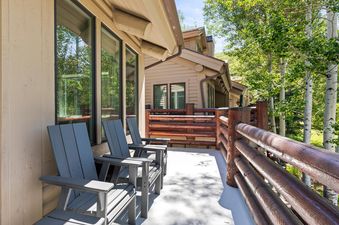 The balcony of Chaparral 204 features comfortable Adirondack chairs and a rustic wooden railing, with lush green trees visible in the background under a bright sky.