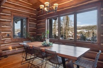 A long wooden dining table with wire mesh chairs in a rustic log cabin dining room with large windows overlooking a snowy mountain landscape.