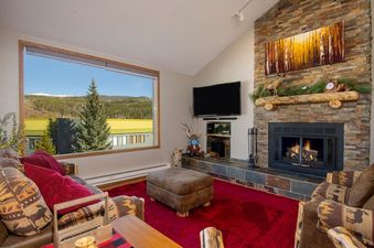 A cozy living room at Snowblaze C24 with a large window offering expansive mountain views, a stone fireplace, and comfortable rustic furnishings.