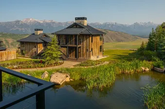 A modern black railing overlooks a serene pond with rustic wooden cabins and majestic snow-capped mountains in the background, creating a picturesque natural landscape.