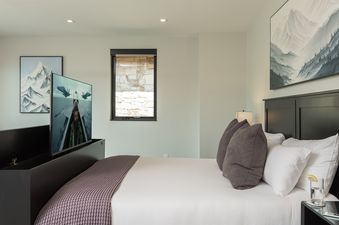 A luxurious bedroom at the Triple 3 Lodge features a large bed, a pop-up TV, and tasteful mountain-themed artwork.