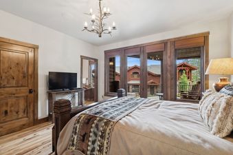 A cozy bedroom at Moose Jaw 34 features a large bed with a patterned throw, a rustic wooden door, and a television, all illuminated by a unique branch-like chandelier and natural light from large windows that offer views of the outdoors.