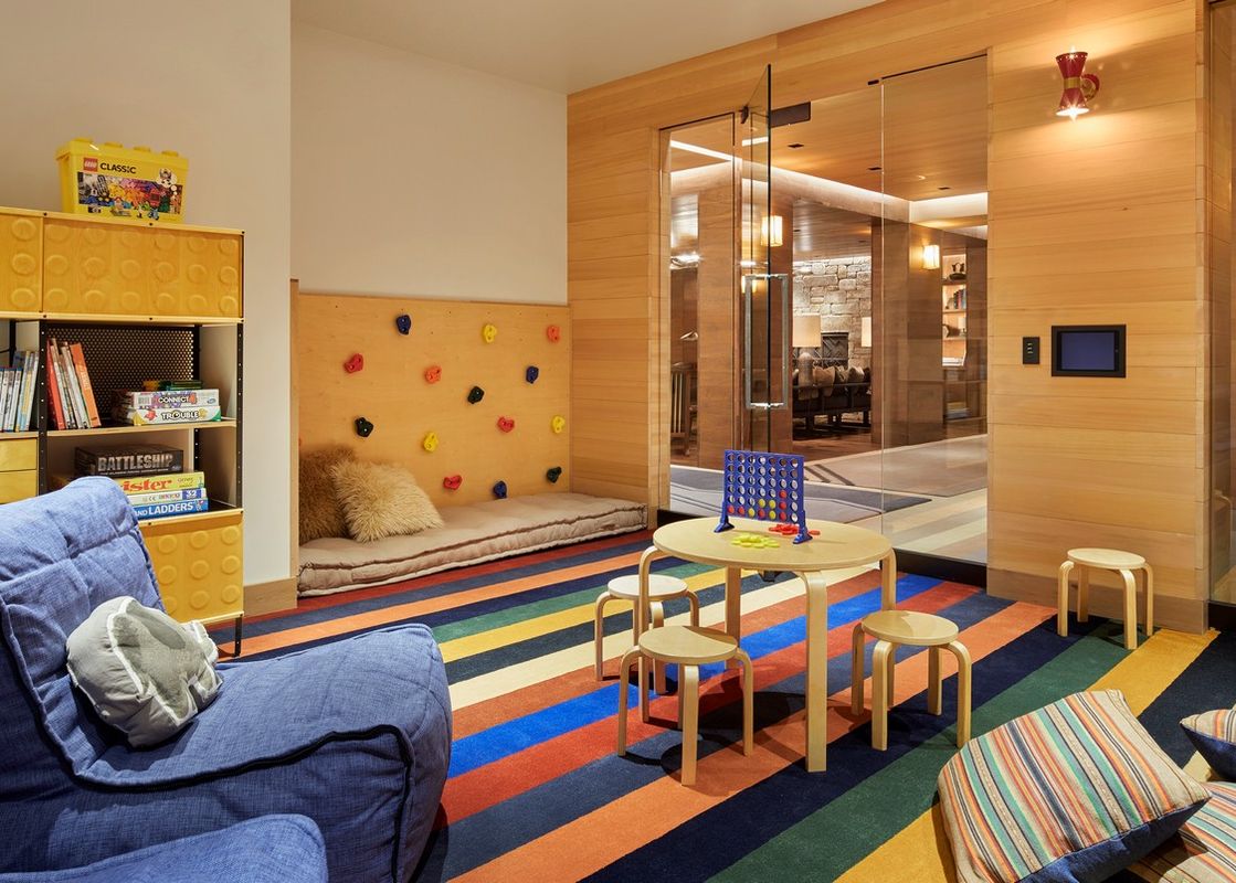 A vibrant and inviting children's play area with a colorful striped rug, a small wooden table with a game of Connect Four, a climbing wall feature, and a LEGO-themed bookshelf.