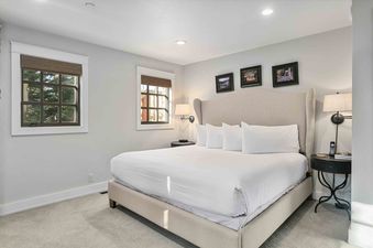A king-sized bed with a tall, upholstered headboard, white linens, and bedside tables with lamps in a cozy bedroom with light gray walls and two windows.