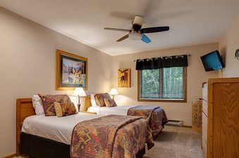 A cozy twin bedroom at Ranch at Steamboat RA401 features two beds with patterned quilts, nature-themed art, a window with outdoor views, a ceiling fan, and a wall-mounted TV.