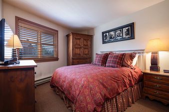 A cozy bedroom at Chateau Chamonix CX121 features a bed with red and gold bedding, wooden furniture, and a window with blinds.