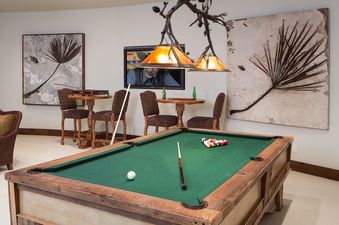 The games room at Phillips Ridge features a billiards table, two high-top tables with chairs, a wall-mounted TV, and unique natural art.