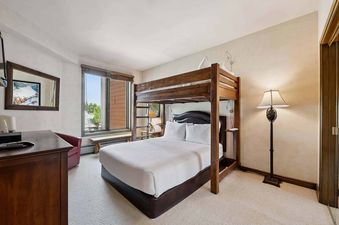 A guest bedroom in Highlands Slopeside 213 features a wooden bunk bed with a full mattress on the bottom and a twin on top, a window, a wooden dresser, a red accent chair, and a floor lamp.