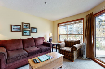 The living area of Aspens on Blackcomb Unit 206 features a comfortable brown sofa, a plaid armchair, and a wooden coffee table, all set on a light-colored carpet.