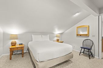 A bright and cozy bedroom in Snow Flower 41 Park City featuring a queen bed with white linens, two wooden nightstands with lamps, a modern digital alarm clock, a slanted ceiling, light-colored carpet, and a wooden chair with a blue cushion beneath a woode