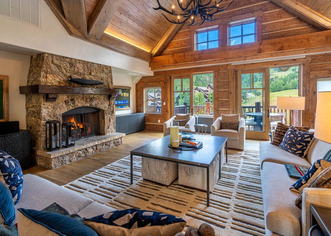 A luxurious living room with a large stone fireplace, plush seating, and expansive windows offering a view of a lush mountain landscape.