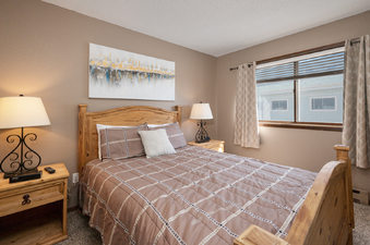 The bedroom at Twin Rivers Vasquez 3 features a queen-sized wooden bed with a brown and white patterned comforter, two bedside tables with lamps, and a window with blinds and curtains.