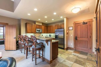 A kitchen with dark wood cabinets, stainless steel appliances, and granite countertops. There are four bar stools, a brown door to the right, and a door with a view of the outdoors to the left.