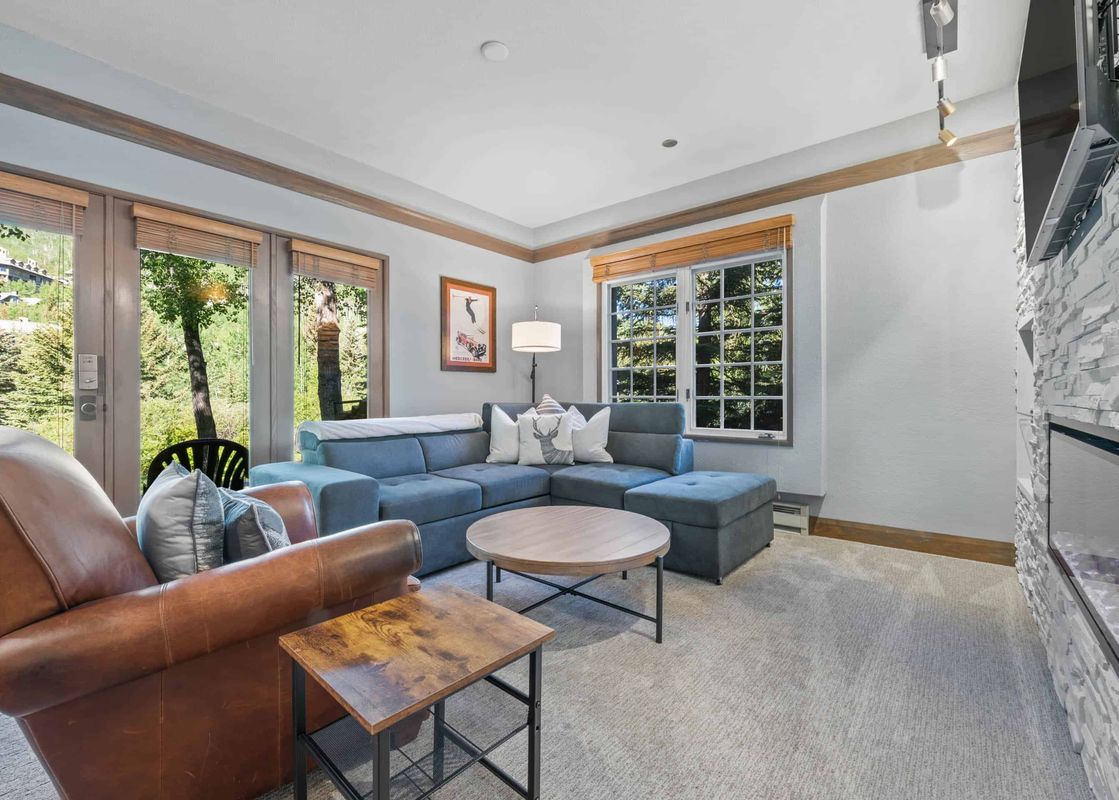 A cozy living room in The Charter E0205 with a blue sectional, leather armchair, wooden tables, and large windows looking out to green trees and mountains.