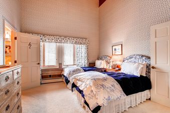 A cozy guest bedroom with two twin beds featuring blue velvet comforters and floral duvet covers, adorned with patterned wallpaper and antique furniture.
