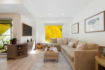 A cozy living room with an L-shaped sofa, an ottoman, and a large window showcasing bright yellow autumn trees outside.
