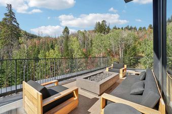 An expansive outdoor balcony at Founders Place 1701, furnished with comfortable lounge seating and a modern fire pit, overlooking a vibrant forest under a clear sky.