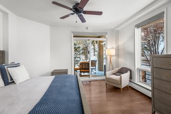 A bedroom with a king-size bed, a dresser, a chair, and a sliding glass door leading to a patio overlooking a snowy forest.