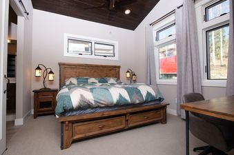 A rustic bedroom with a wooden bed, patterned bedding, and large windows looking outside.