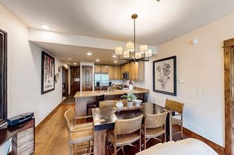 Dining area with a wooden table, woven chairs, and a chandelier, with the kitchen in the background in BlueSky Breckenridge 402.