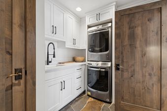 A functional laundry room featuring white cabinetry, a utility sink with a black faucet, and a stacked washer and dryer unit, all set against a rustic flagstone floor.