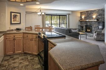 A wide-angle view of the kitchen and living area of Building 4 Two Bedroom Breckenridge Suite, featuring granite countertops, wood cabinets, a dining table, a stone fireplace, and large windows with a snowy forest view.