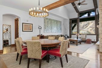 A dining area with a round wooden table and six chairs with red seats and leopard-print backs, set on a rug in an open-plan room with vaulted ceilings and large windows overlooking a snowy mountain landscape.