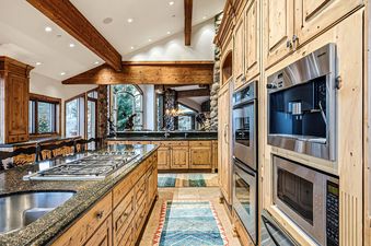 A spacious and luxurious gourmet kitchen with a large island, stainless steel appliances, and rich wooden cabinetry is illuminated by natural light from expansive windows.
