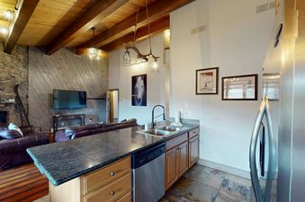 The kitchen in Phoenix 226 has a large island with a double sink, black faucet, and granite countertop, with a view of the living area with a fireplace and television.