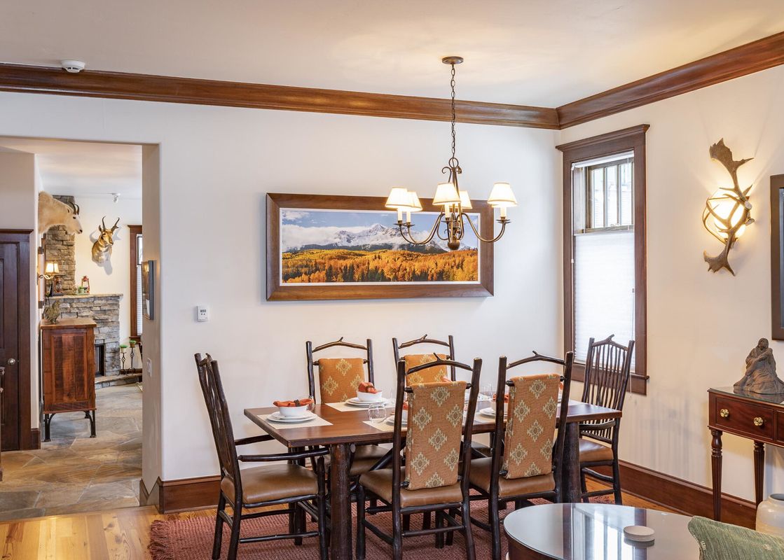 A rustic dining room with a wooden table and chairs, a chandelier, a framed picture of a mountain scene, and a fireplace in the background.