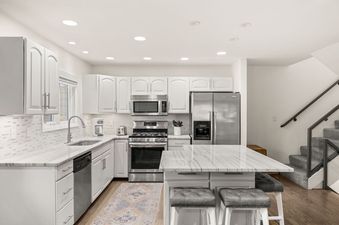 A modern kitchen with white cabinets, stainless steel appliances, marble countertops, and a kitchen island with bar stools.