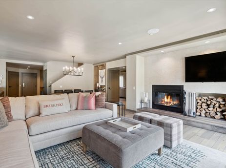 A wide view of the comfortable and stylish living room in Village Hall 09, featuring a large sectional sofa, a fireplace with a TV mounted above, and a dining area visible in the background.