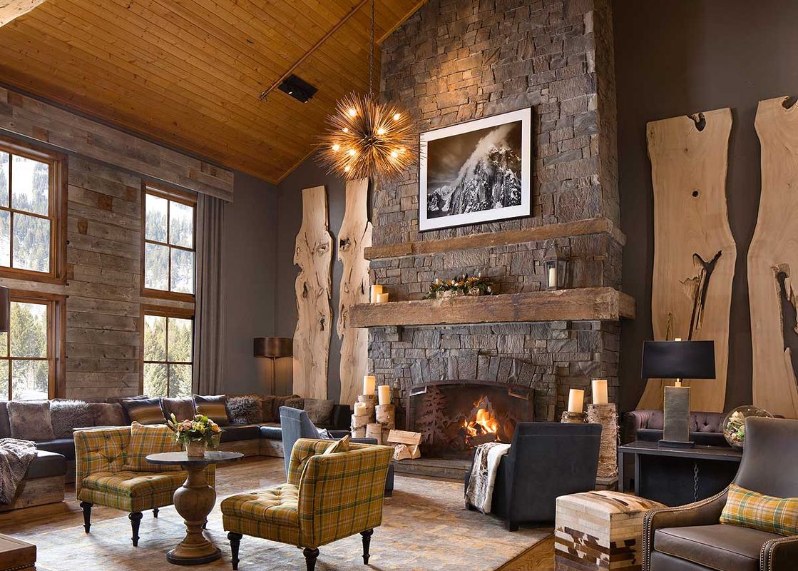 A cozy, mountain-style lounge featuring a grand stone fireplace, vaulted wooden ceilings, and plaid armchairs.