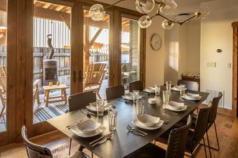 A modern dining room with a large dark wood table set for dinner, featuring contemporary light fixtures and glass doors leading to a bright outdoor patio.